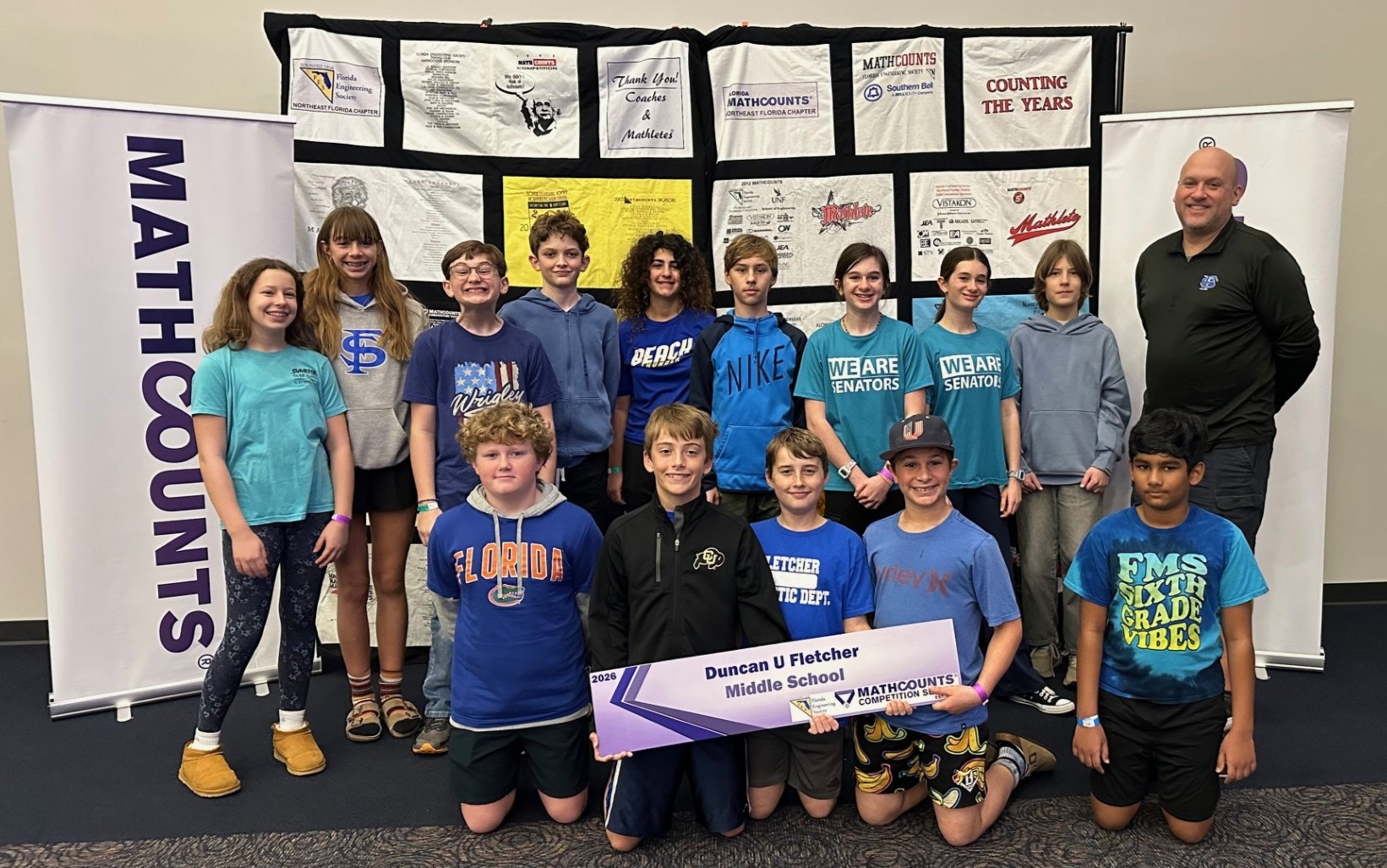 Group of children and adults posing with a large check in a room with Mathcounts banners.