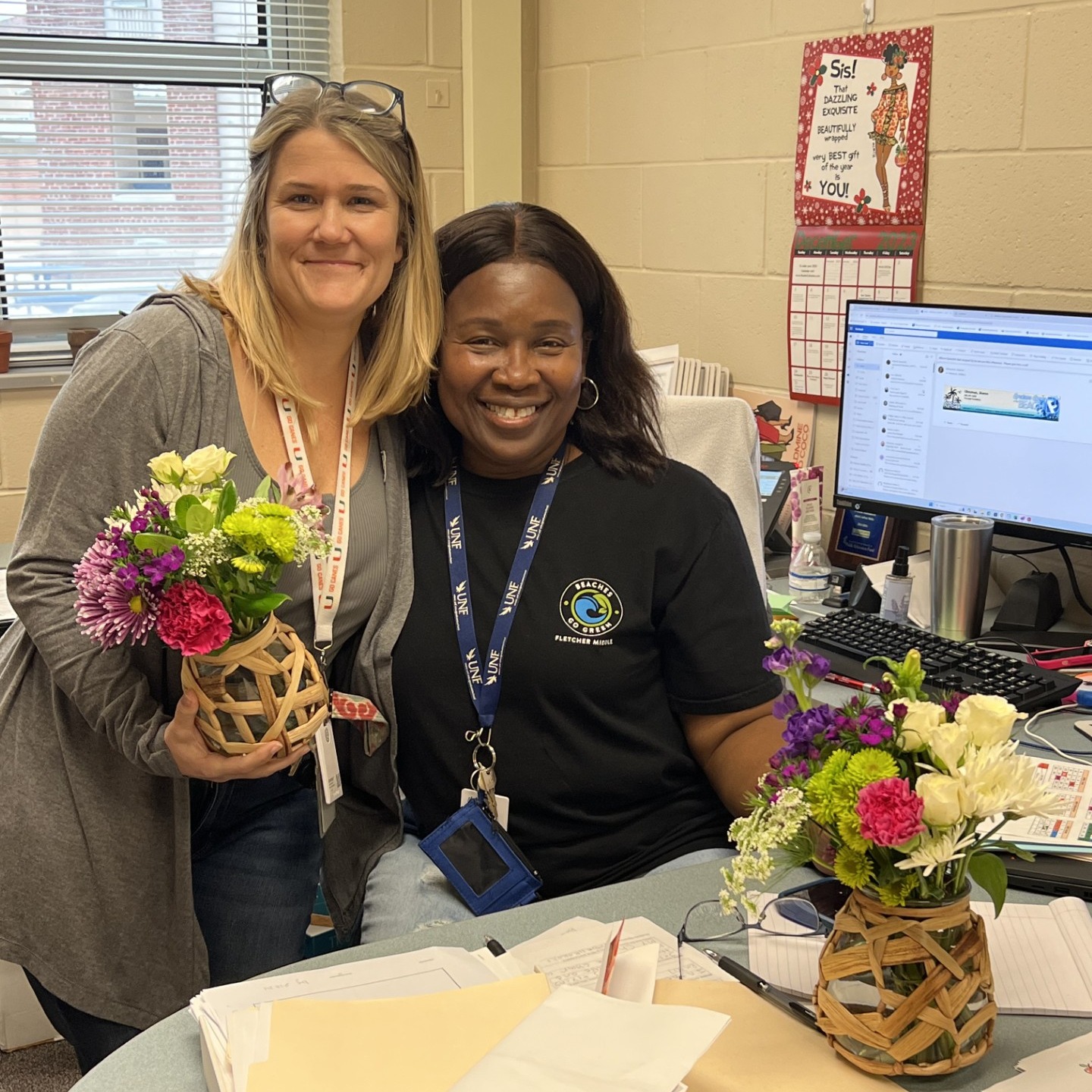 Two women smile in front of a desk with flowers, a computer monitor, keyboard, and papers.