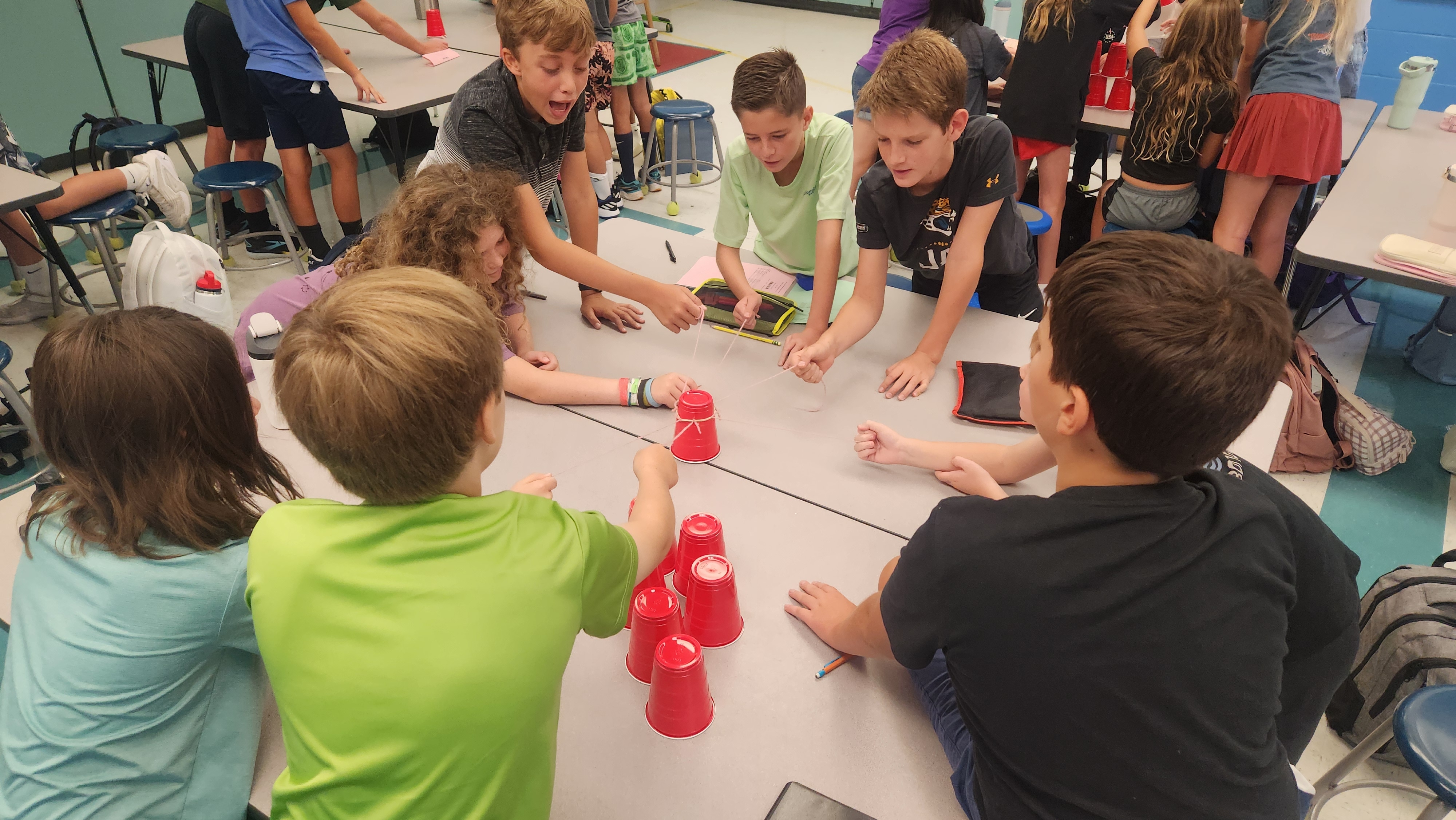 Children seated around a table playing a game using red plastic cups in a classroom.
