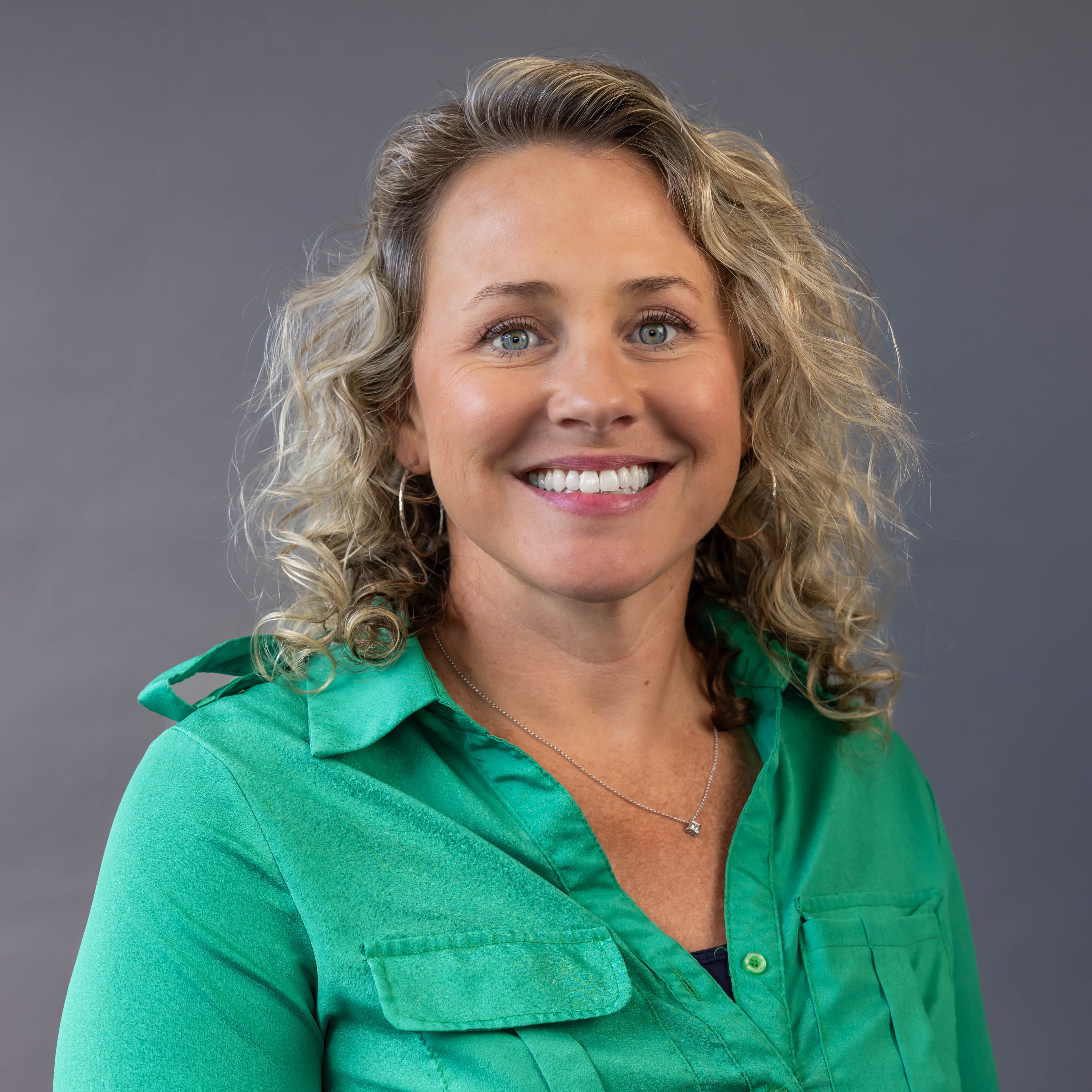 A woman with blonde curls wearing a green blouse, smiling, and standing against a gray background.