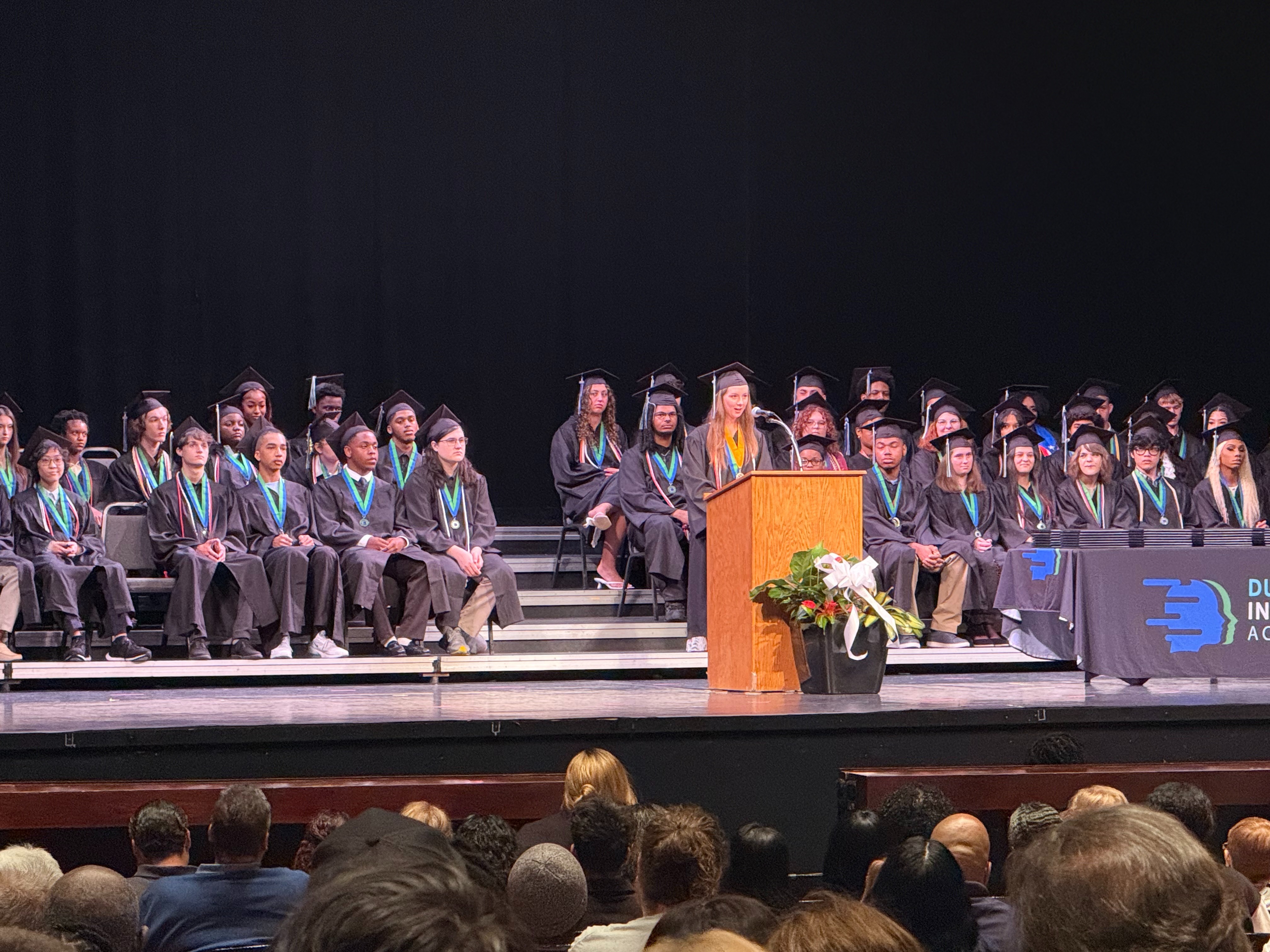 Graduates in caps and gowns seated on a stage. A podium with flowers is in the middle. An audience is watching.