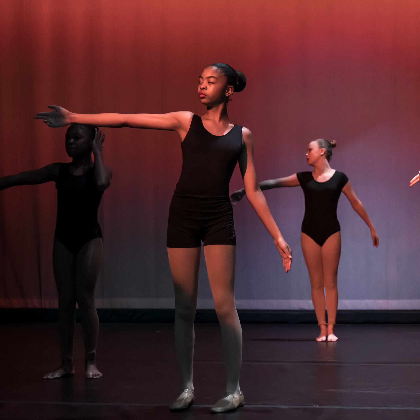 Four dancers in black attire perform on a stage with a red background, executing ballet moves and poses.