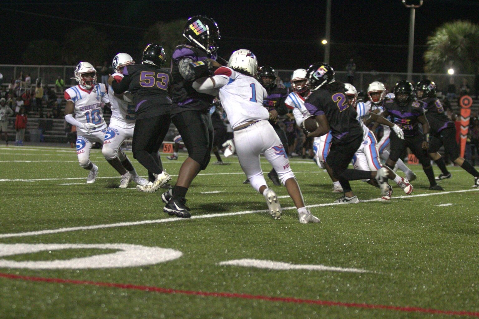 Football players in action on a field, with numbers on jerseys. Players wear helmets, gloves, and sneakers.