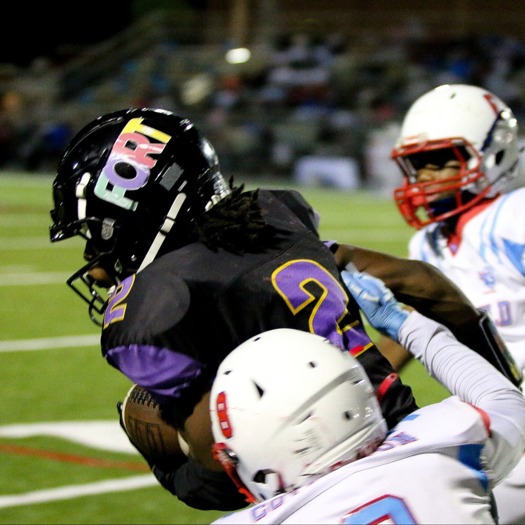 Four football players engage on a field. Two wear white helmets, one in purple and one in blue.