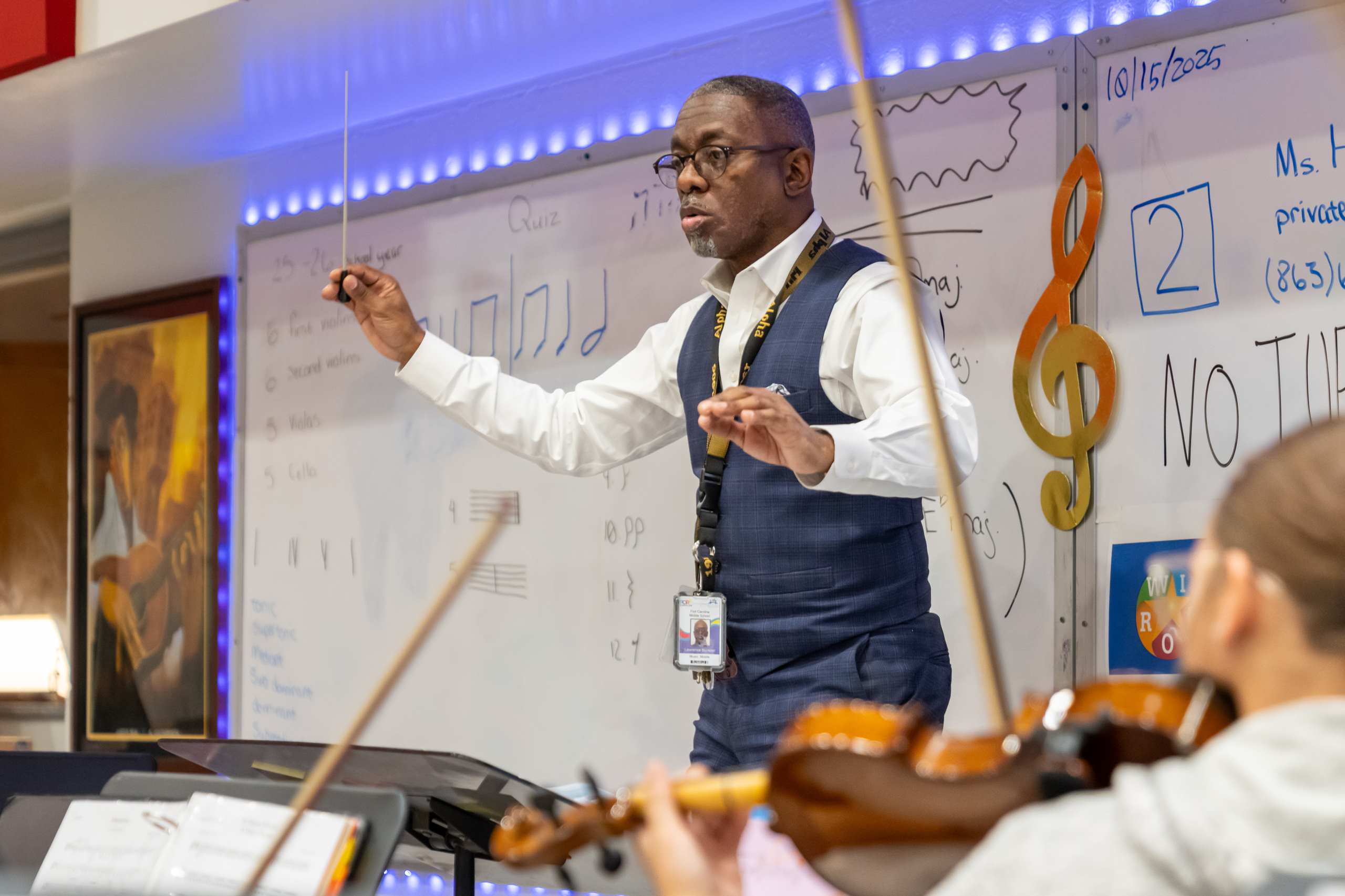 A man directs a violin ensemble; a student plays a violin while facing a whiteboard with musical notations.
