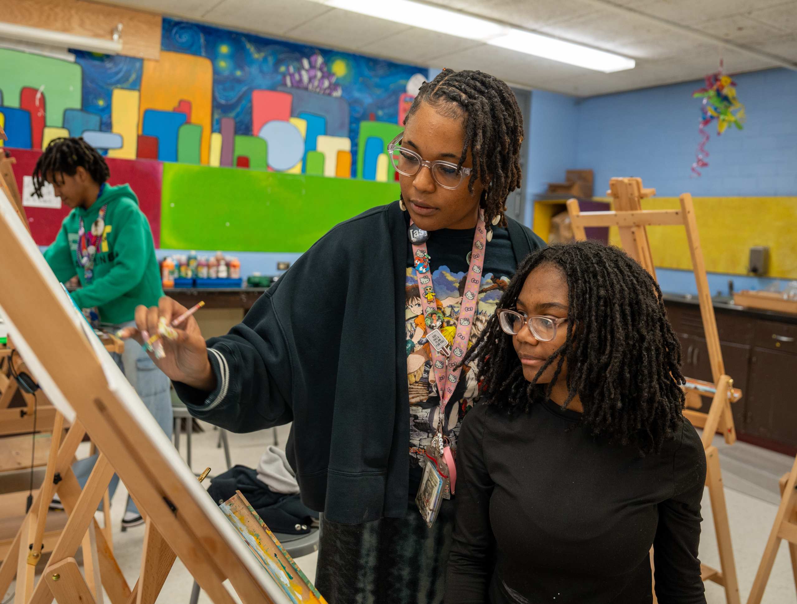 An adult and a child stand before easels, both wearing glasses. A colorful mural adorns the background.