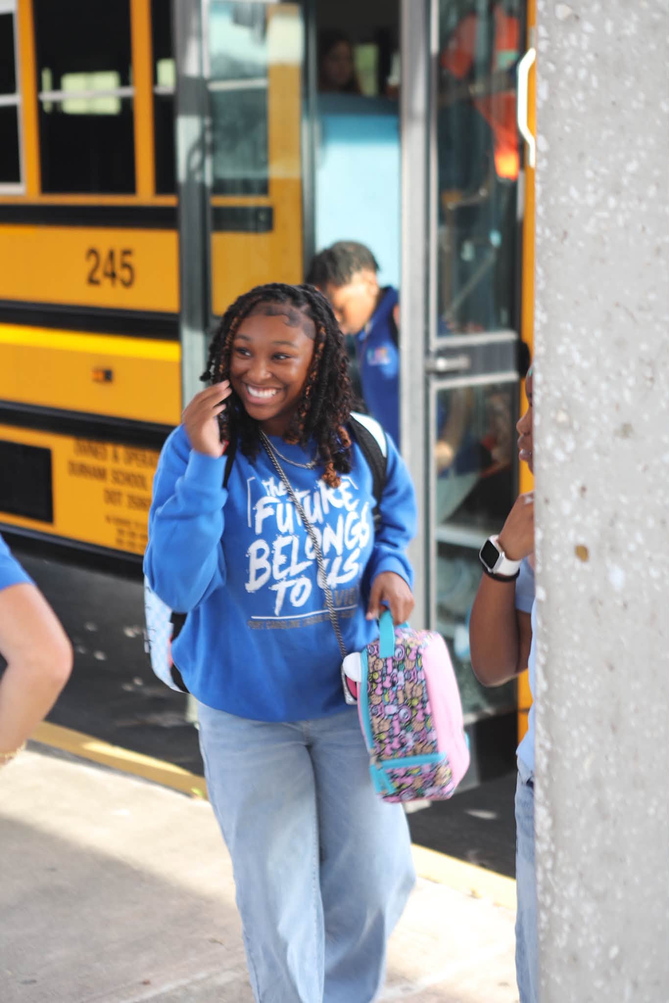 A woman in a blue sweatshirt and jeans, smiling and talking on her phone, stands in front of a yellow school bus.