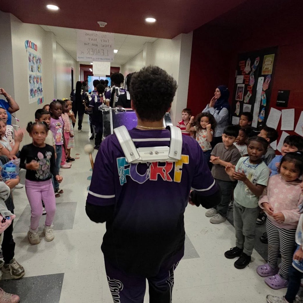Person in a purple shirt walks through a hallway with children standing in line, red walls, and overhead lights.