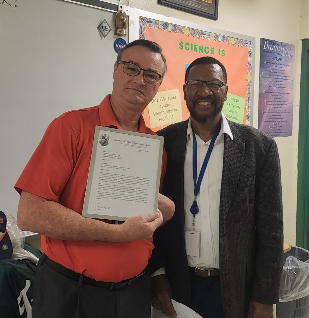 Two men in a classroom. One holds a framed certificate. A bulletin board with colorful posters and text is behind them.