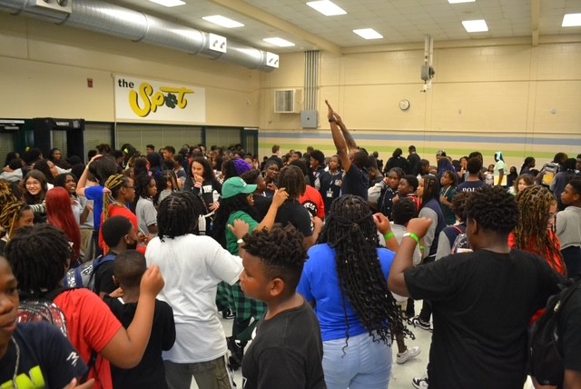 A large crowd of people in a room, some wearing bracelets, with bright fluorescent lights overhead.