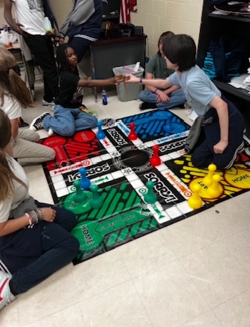 Students sit on floor playing a board game. One student hands another a game piece.