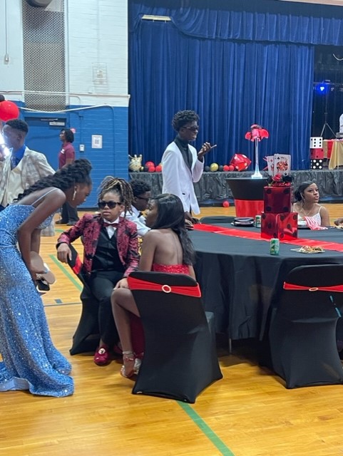 A group of people gathered indoors. A woman in a blue dress speaks to a seated woman in red.