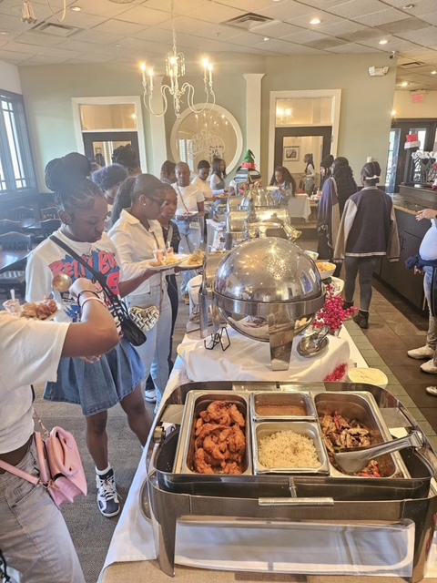 Multiple people stand around a long table with trays of food. Some hold plates. A woman takes a photo.