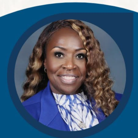 Woman with curly hair, wearing a blue blazer and a white collared top, smiling. The background is blue with a circular pattern.