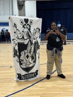 Person in black shirt and khaki pants stands next to a banner with a crest on a basketball court.