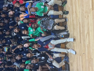 A group of students in green shirts sit on bleachers in a gymnasium. Many more students fill the seats behind them.