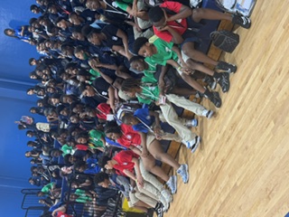 A gym with a crowd of students in various colored shirts sitting on bleachers, listening.