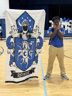 Person in blue shirt and khaki pants standing next to a white banner with a crest.