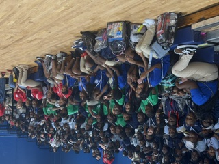 People sitting on bleachers in a gymnasium. Some have backpacks. A crowd is seated in the stands behind them.