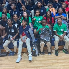 Group of young students wearing green shirts and ID lanyards seated on bleachers, some holding bags.
