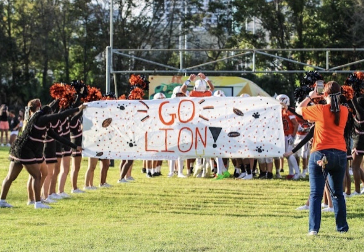 Cheerleaders hold a banner that reads "GO LION!" on a grassy field, with trees and a metal fence in the background.