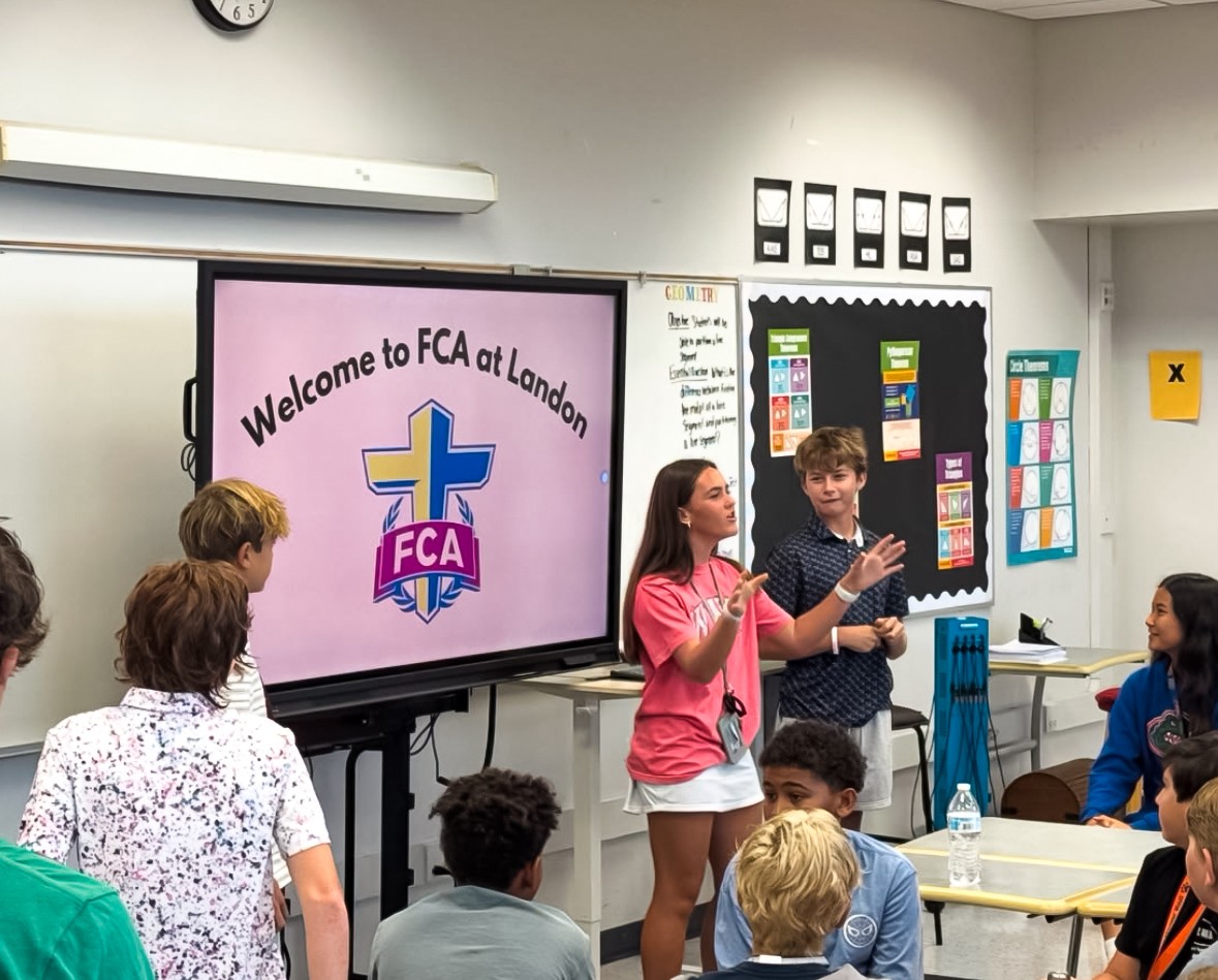 A group of students in a classroom, with one presenting in front of a large screen with a welcome message.