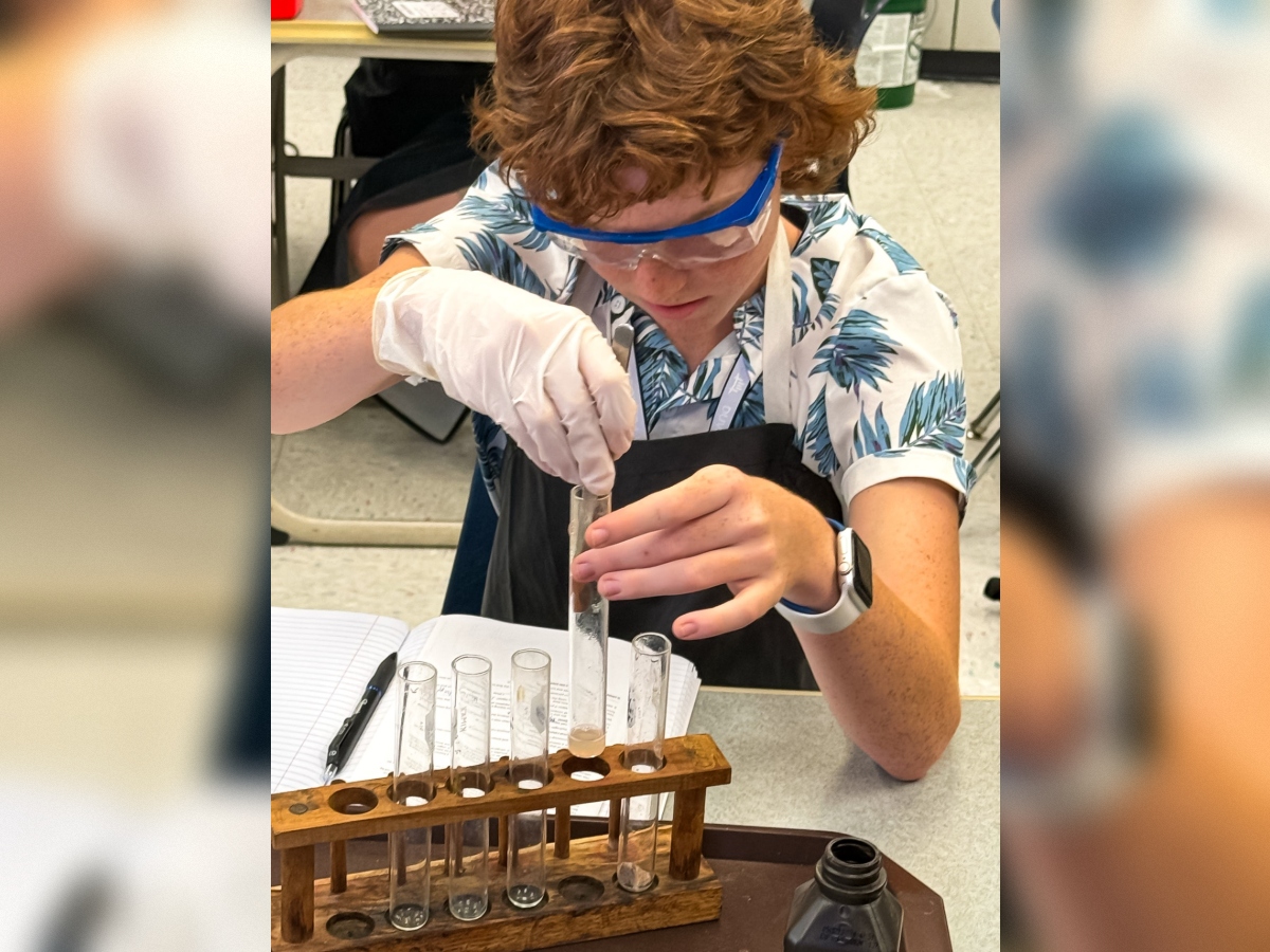 A person in a lab coat and goggles conducts an experiment, pouring liquid into a test tube.