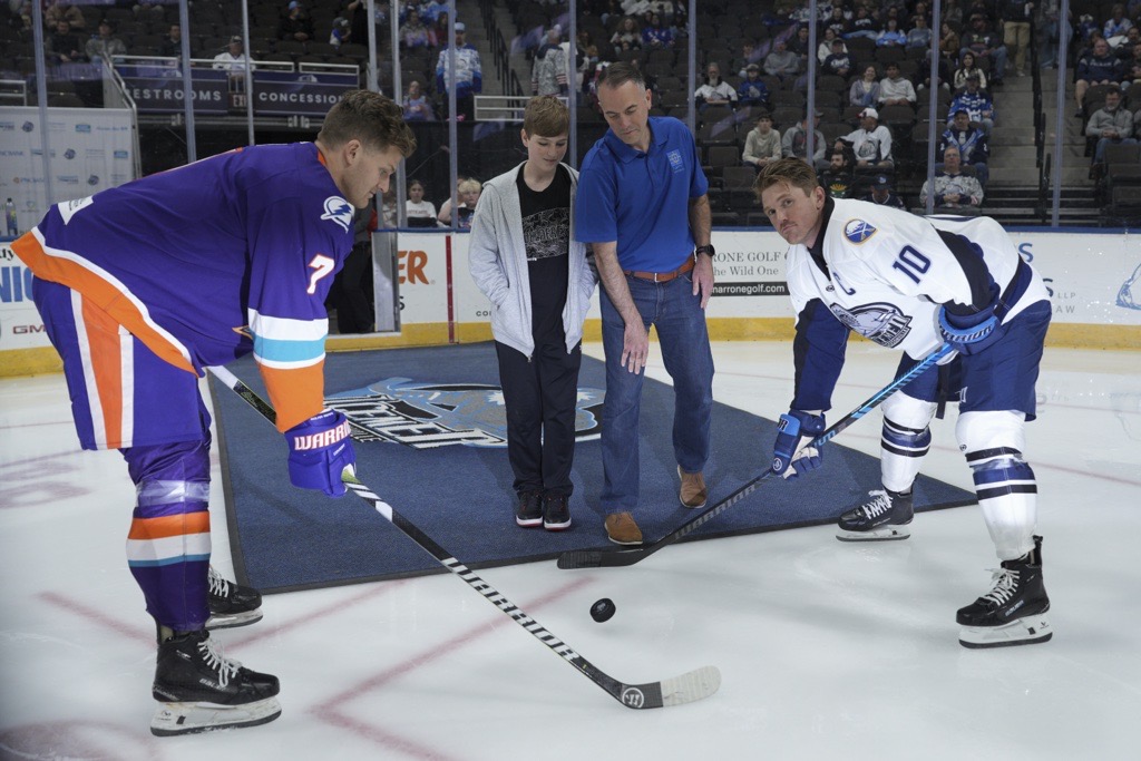 Three hockey players in jerseys, two holding sticks, play on ice. Spectators stand nearby. Audience sits in stands at rear.