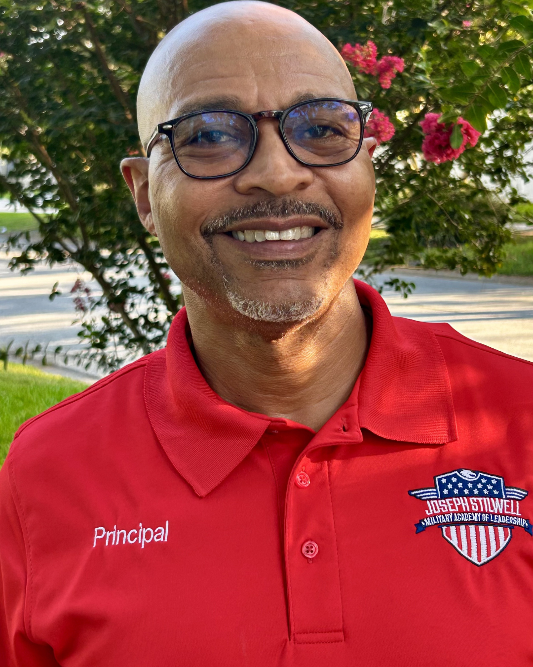 Person in red polo shirt with "Principal" and logo. Bald, wearing glasses, and smiling. Blurry background with greenery.