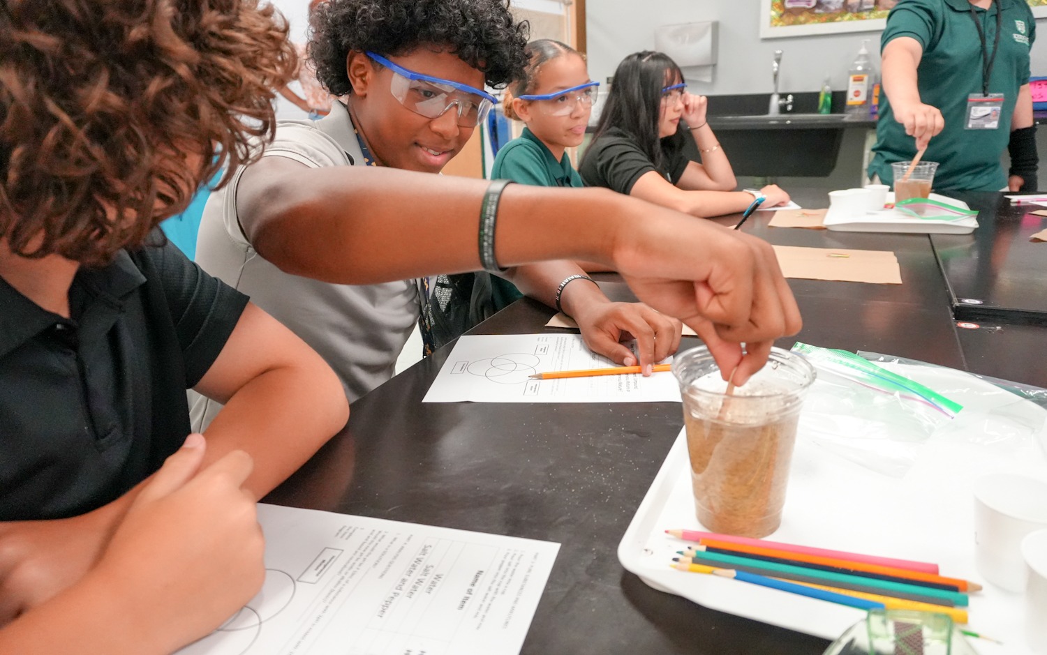 Students seated at a table in a classroom, wearing safety goggles. One student is adding a substance to a cup.