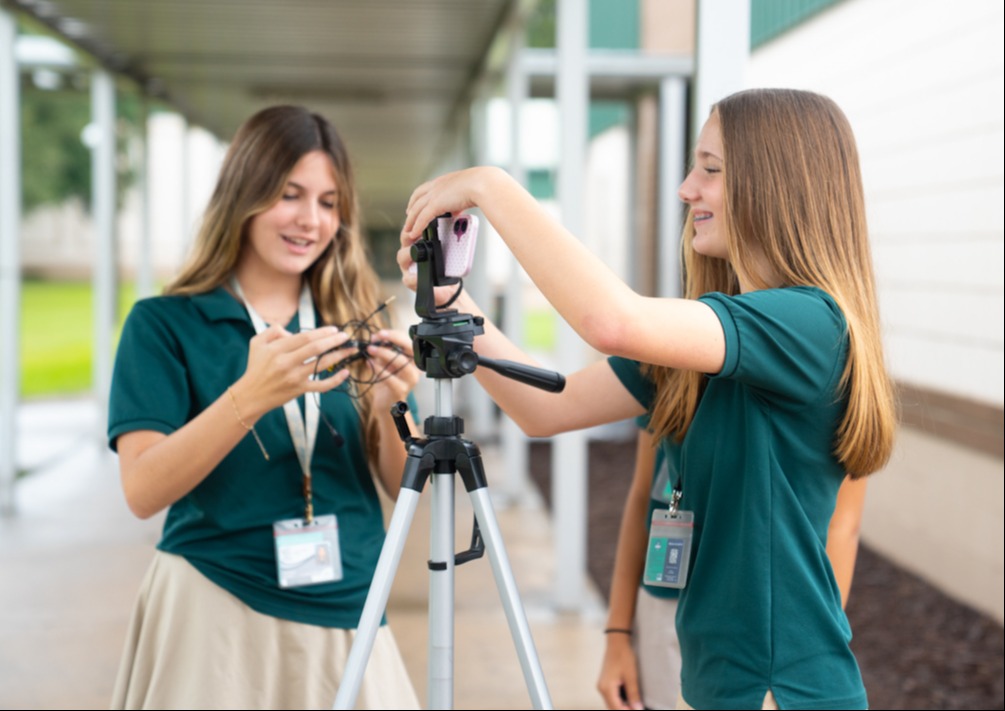 Two women with green tops and ID cards set up a tripod and camera in an outdoor school area.