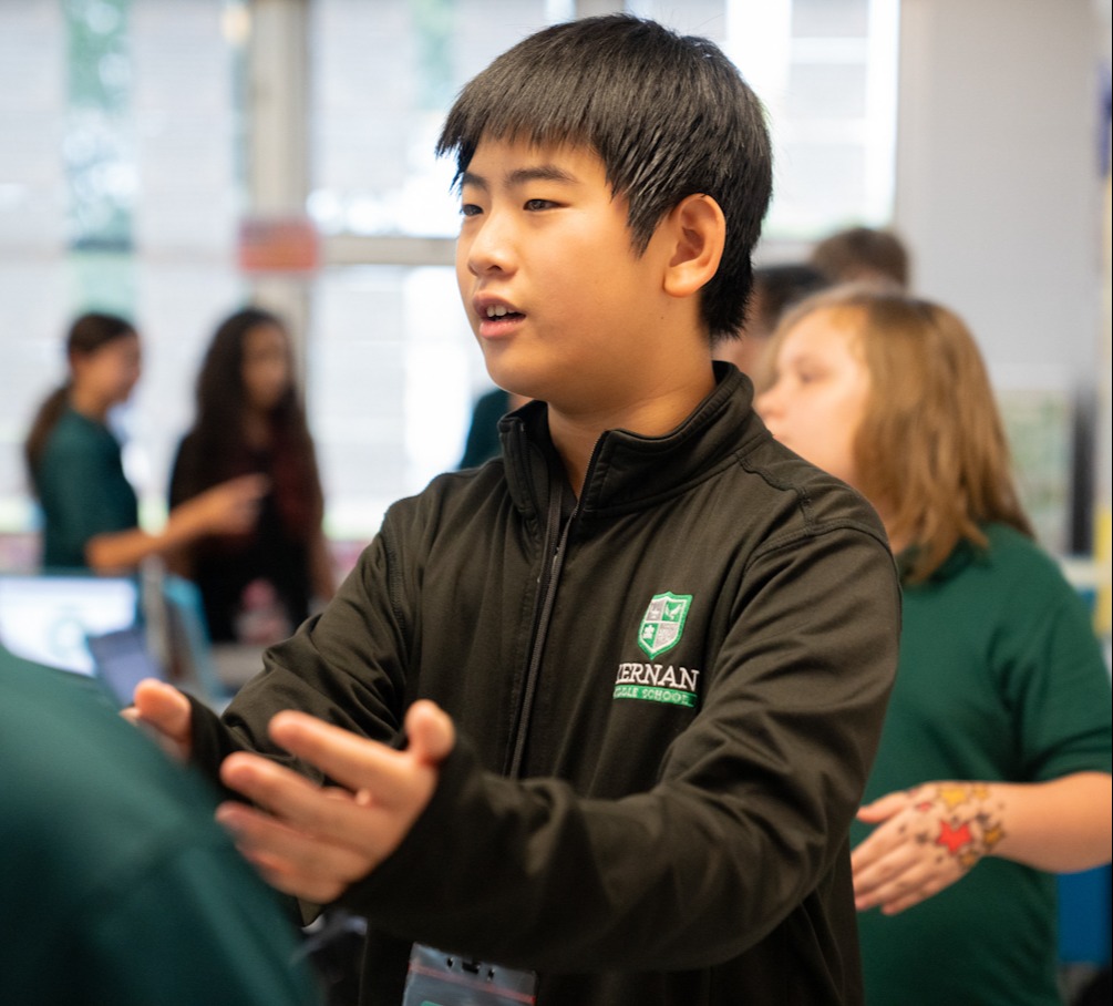 A young boy with short hair in a black jacket speaks to another student. Behind him, two girls and two adults stand.