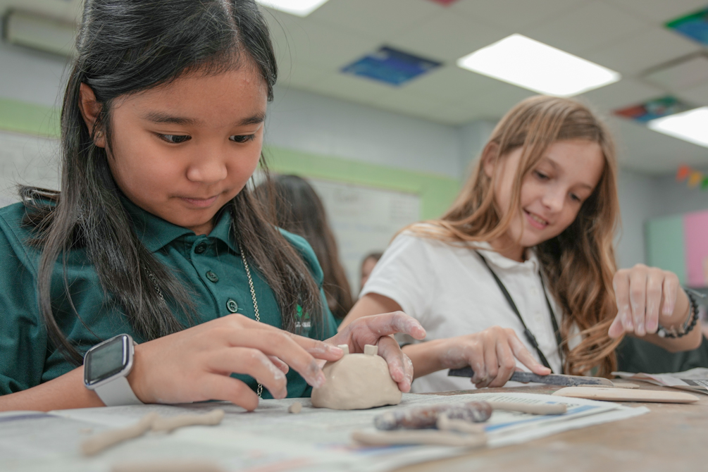 Two girls in a classroom, one with long hair molding clay while the other holds clay. Table with clay and objects.