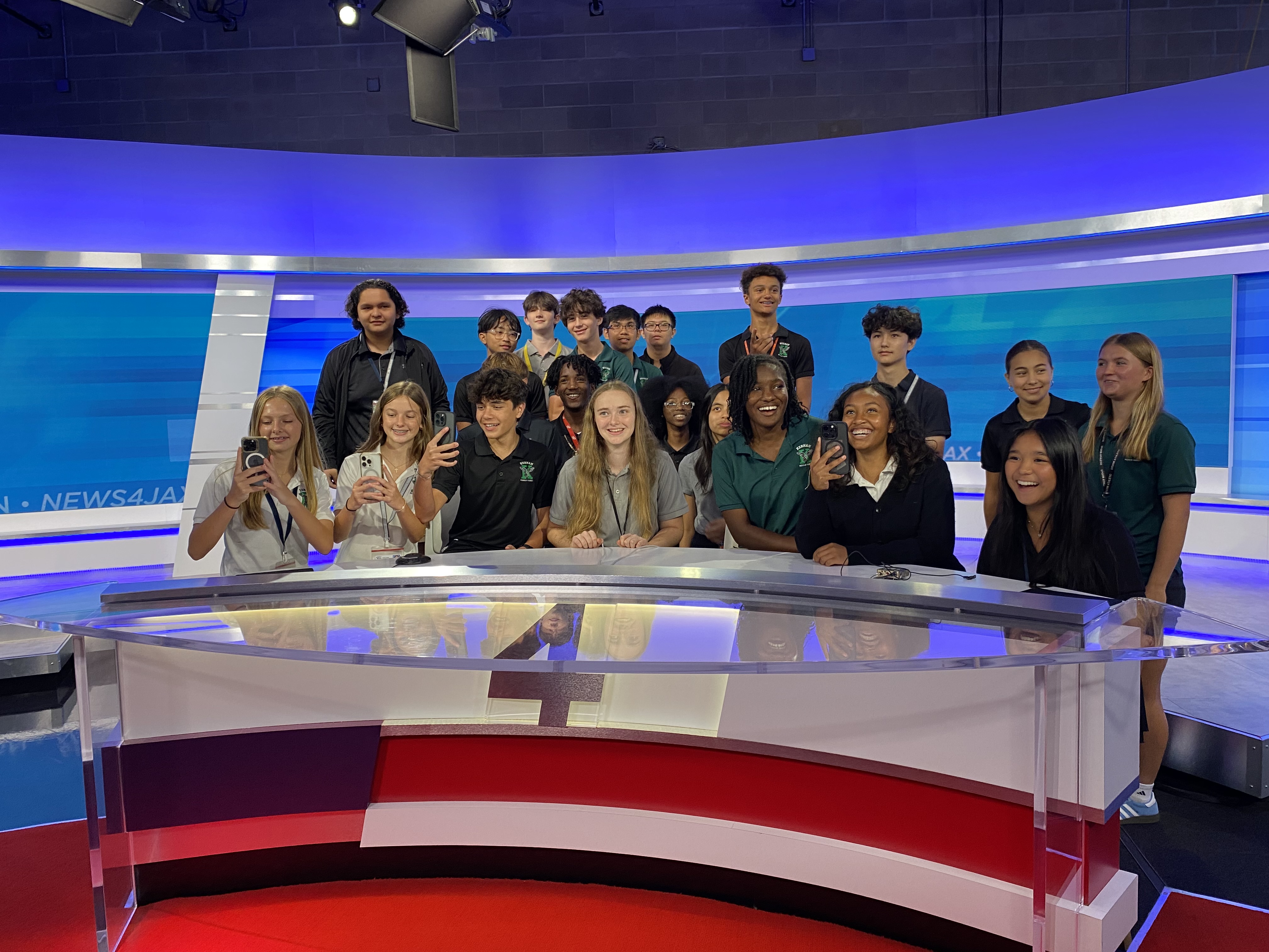 A group of young people, standing behind a news desk, take photos and smile in a studio.
