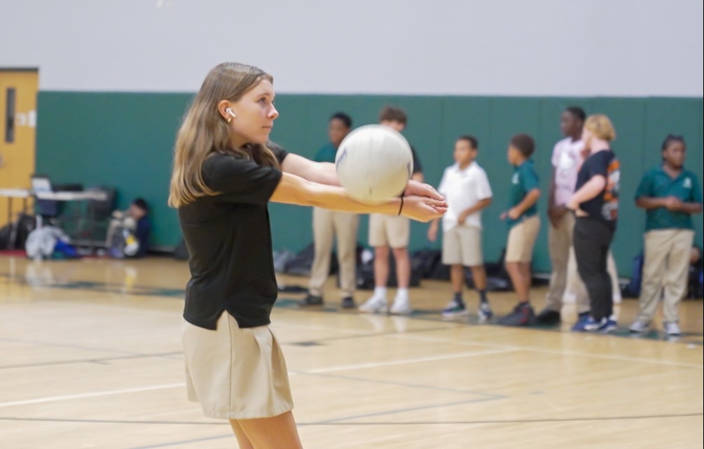 A girl in black top and skirt holds a ball ready to throw. Several people stand behind her on a basketball court.