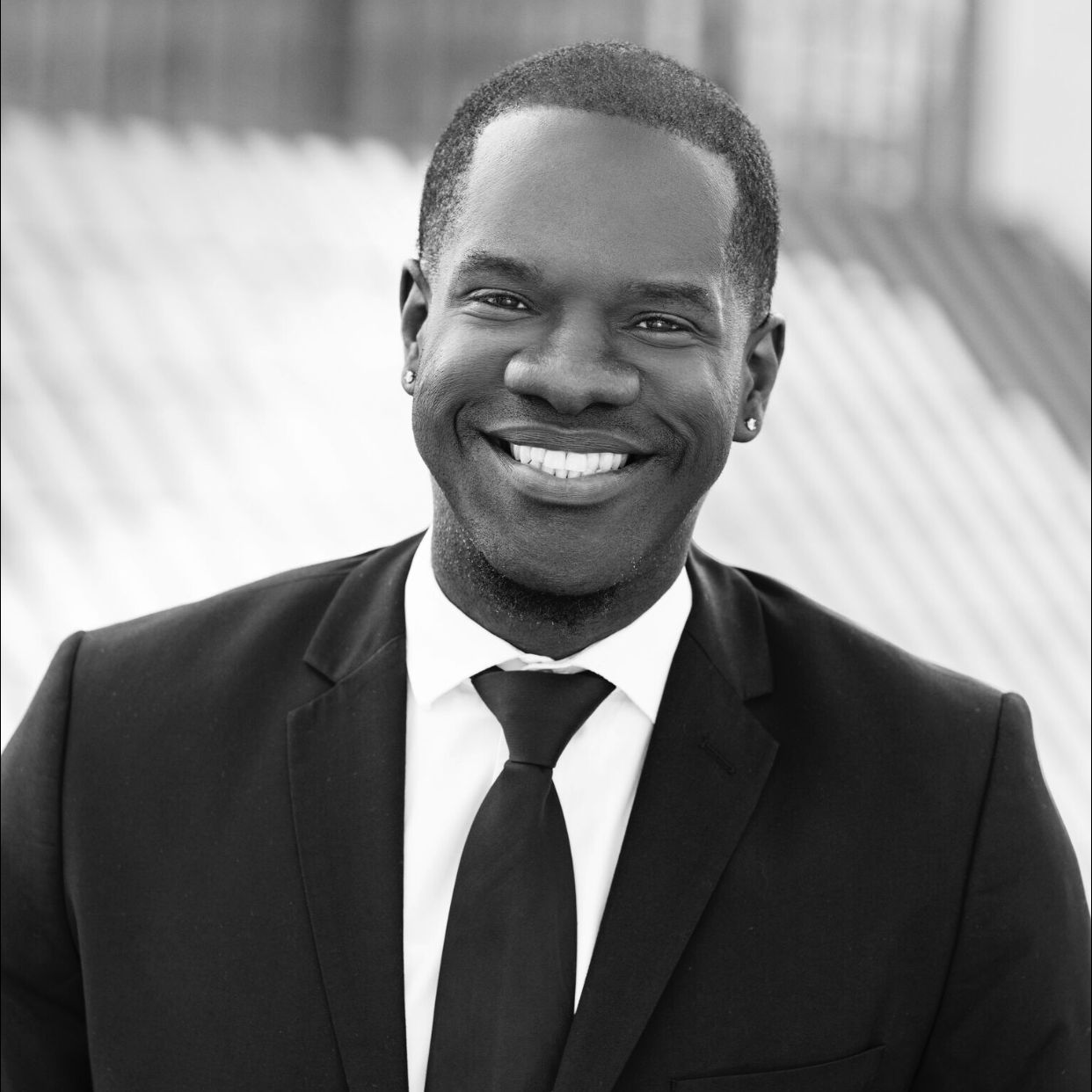 A black-and-white photo of a man smiling, wearing a suit and tie with a patterned background.