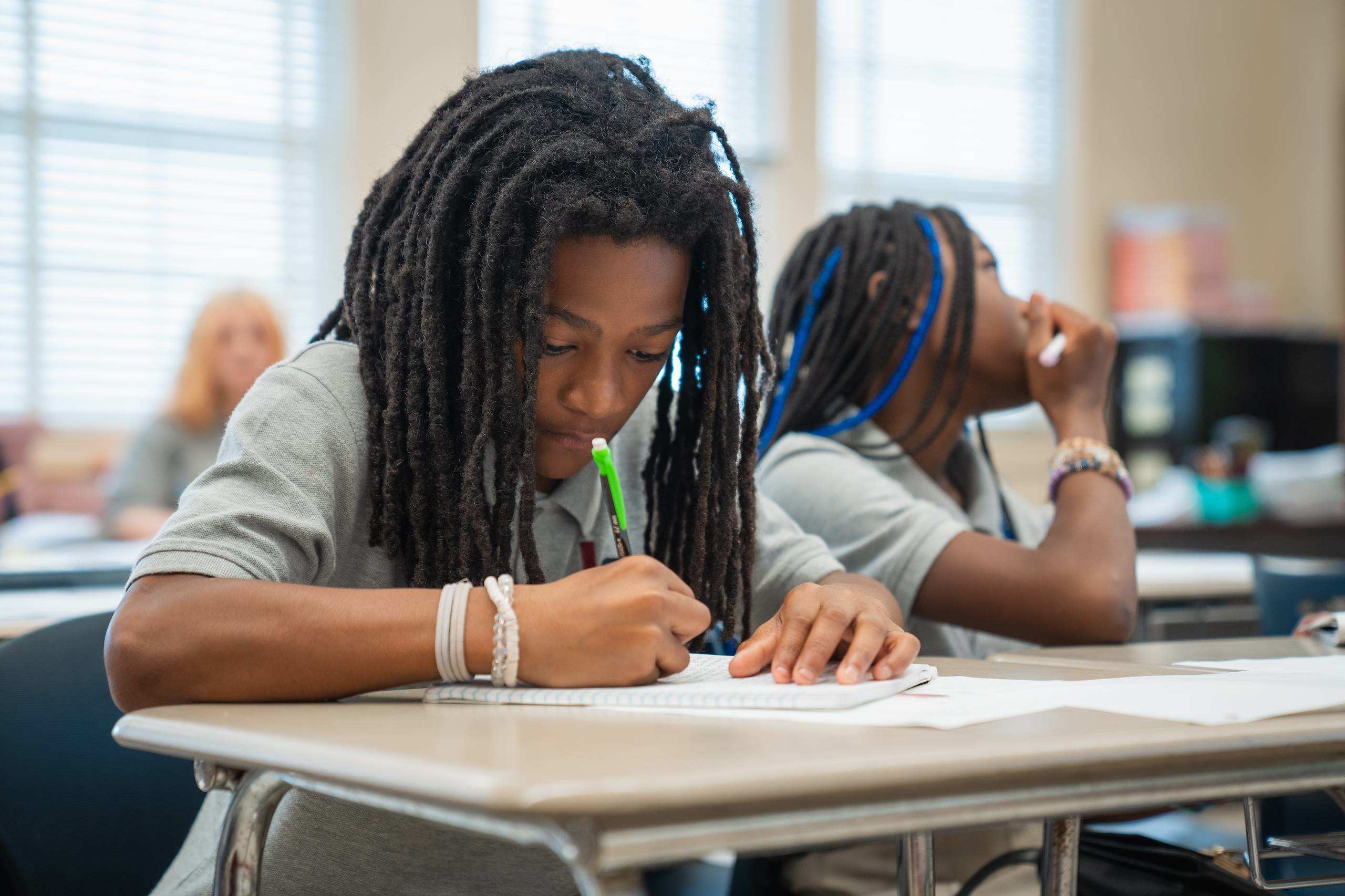 Two students in a classroom, one writing on paper, another with hands on their face.