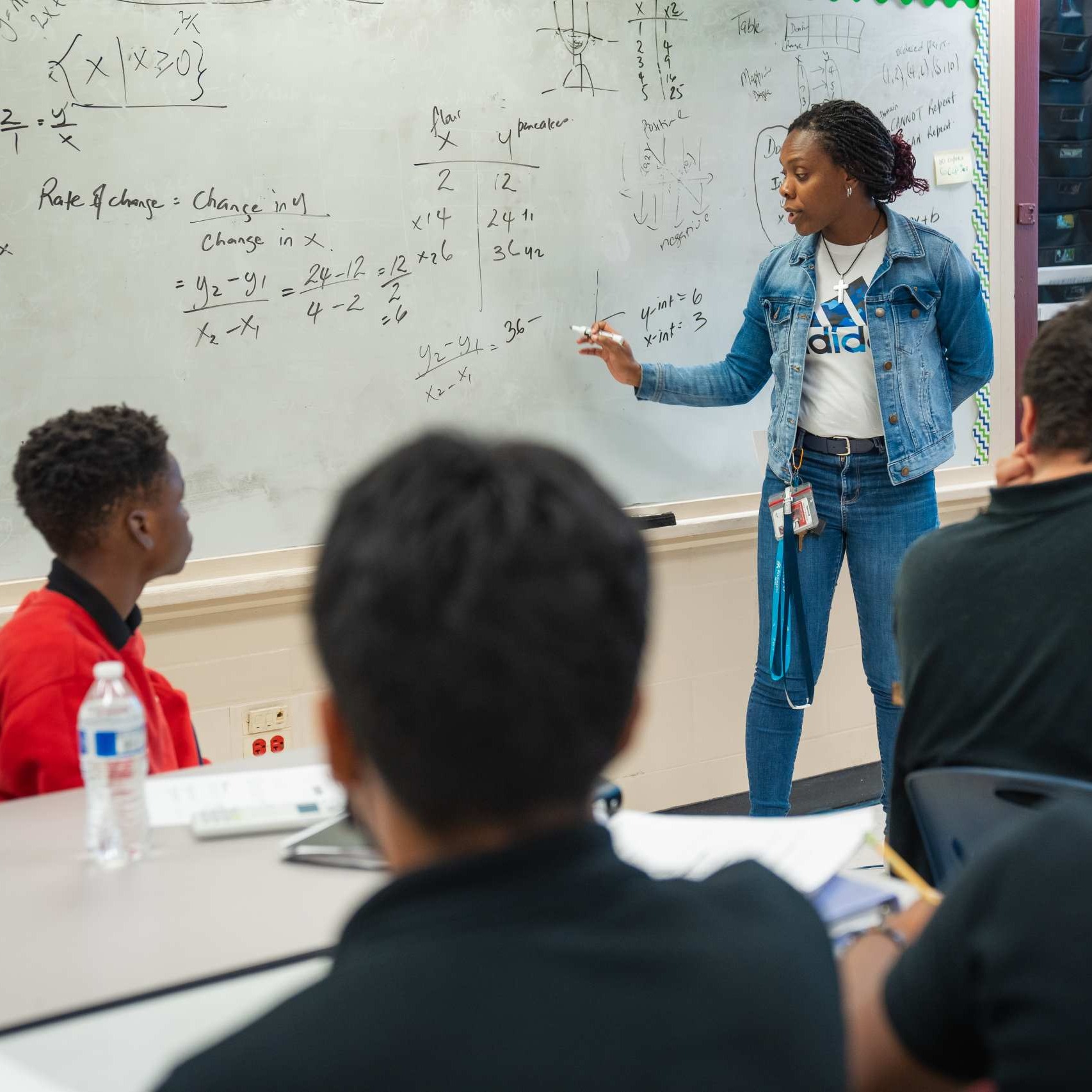 A teacher, wearing a blue jacket and jeans, stands in front of a whiteboard with mathematical formulas. Students sit at a table, facing the board.