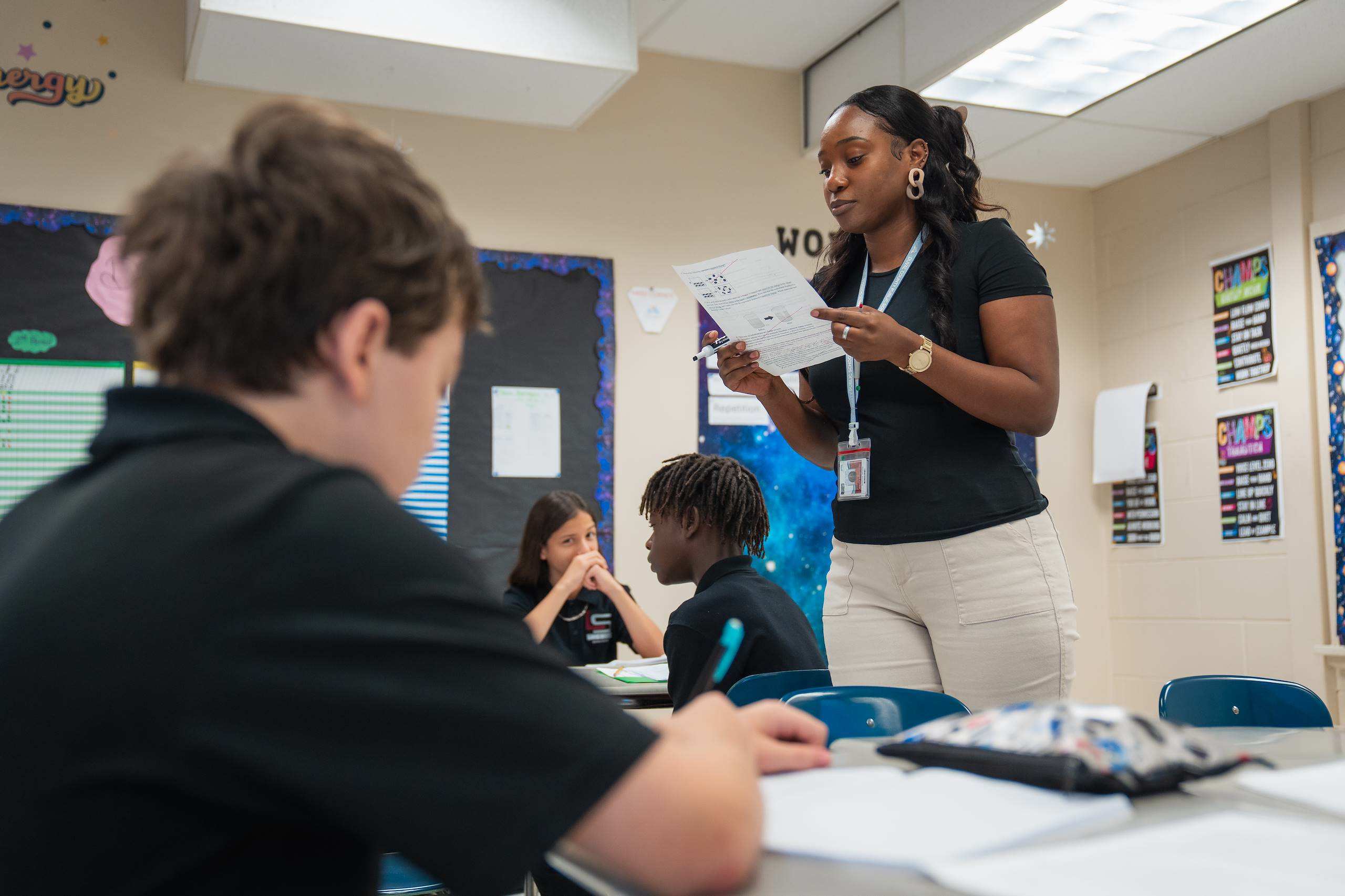 Teacher stands at a table reading a paper to a student. Other students are seated at desks behind them.