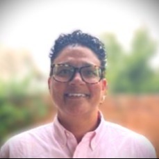 Photograph of an individual in a pink shirt, wearing glasses with a short haircut.  The background is blurry.  The photograph is taken from the shoulders up.