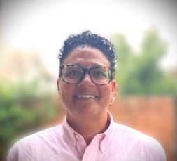 Person with short curly hair, glasses, and a pink shirt, smiling and standing against a blurred green background.