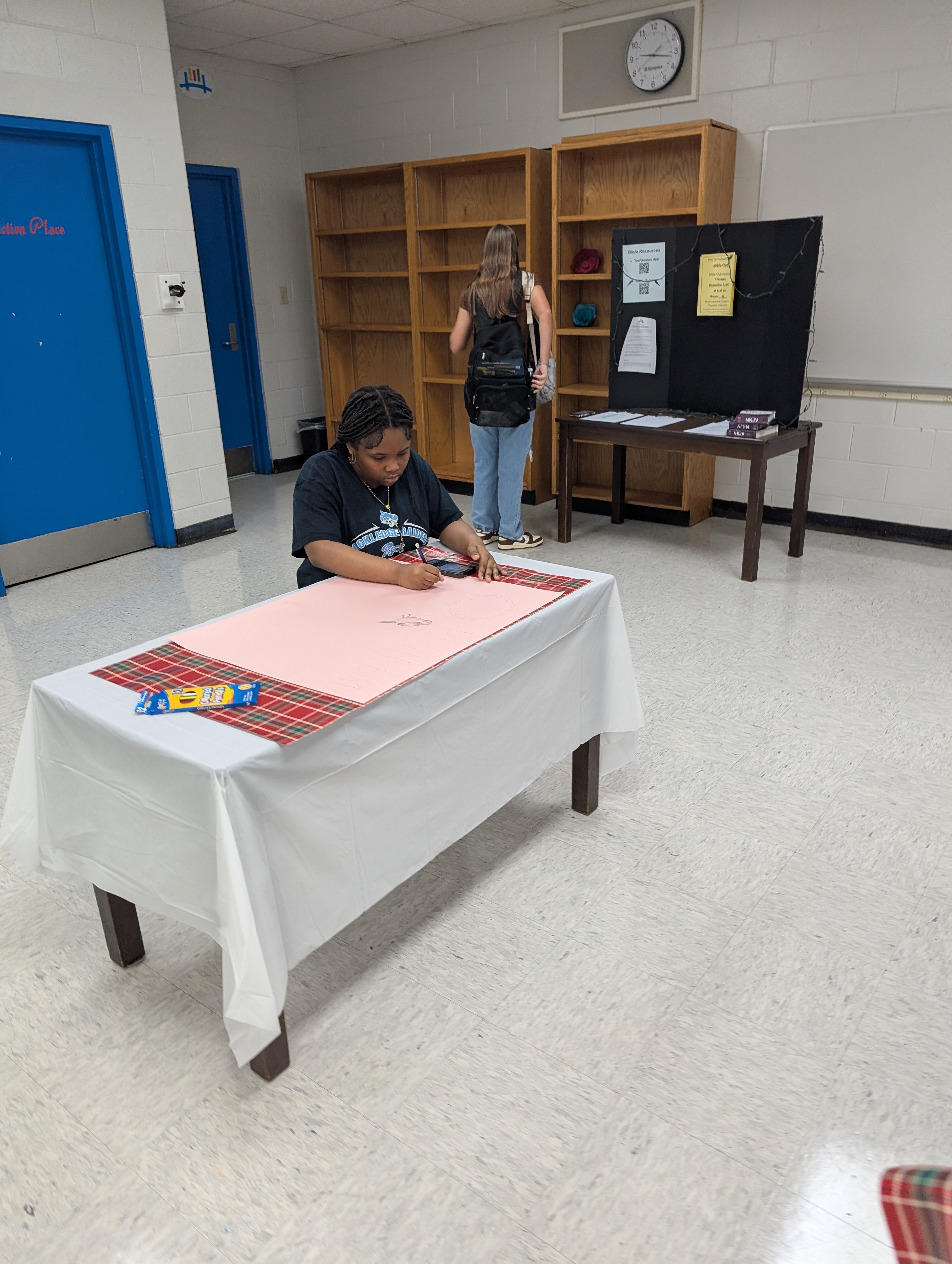 A woman writes on a pink sheet on a covered table. A blue door is on the left, and a woman in the background walks by a bulletin board.