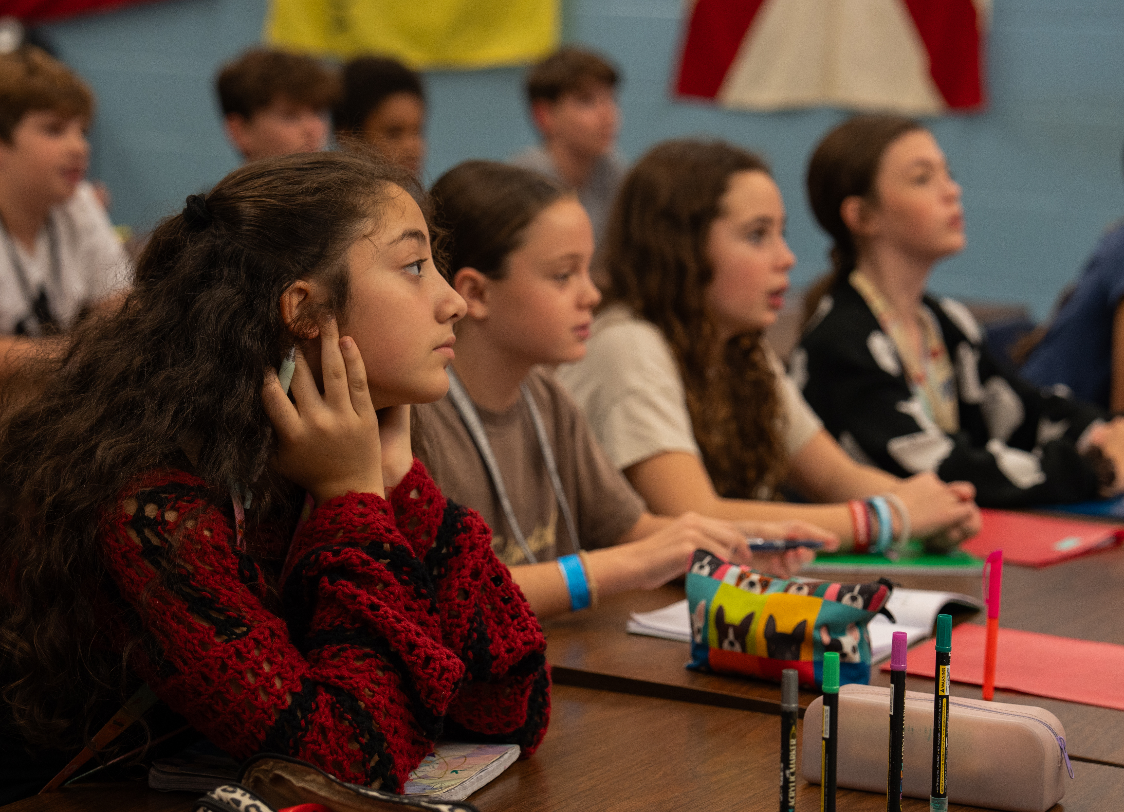 Several students seated in a classroom. One girl in a red top is looking down and holding her ear.