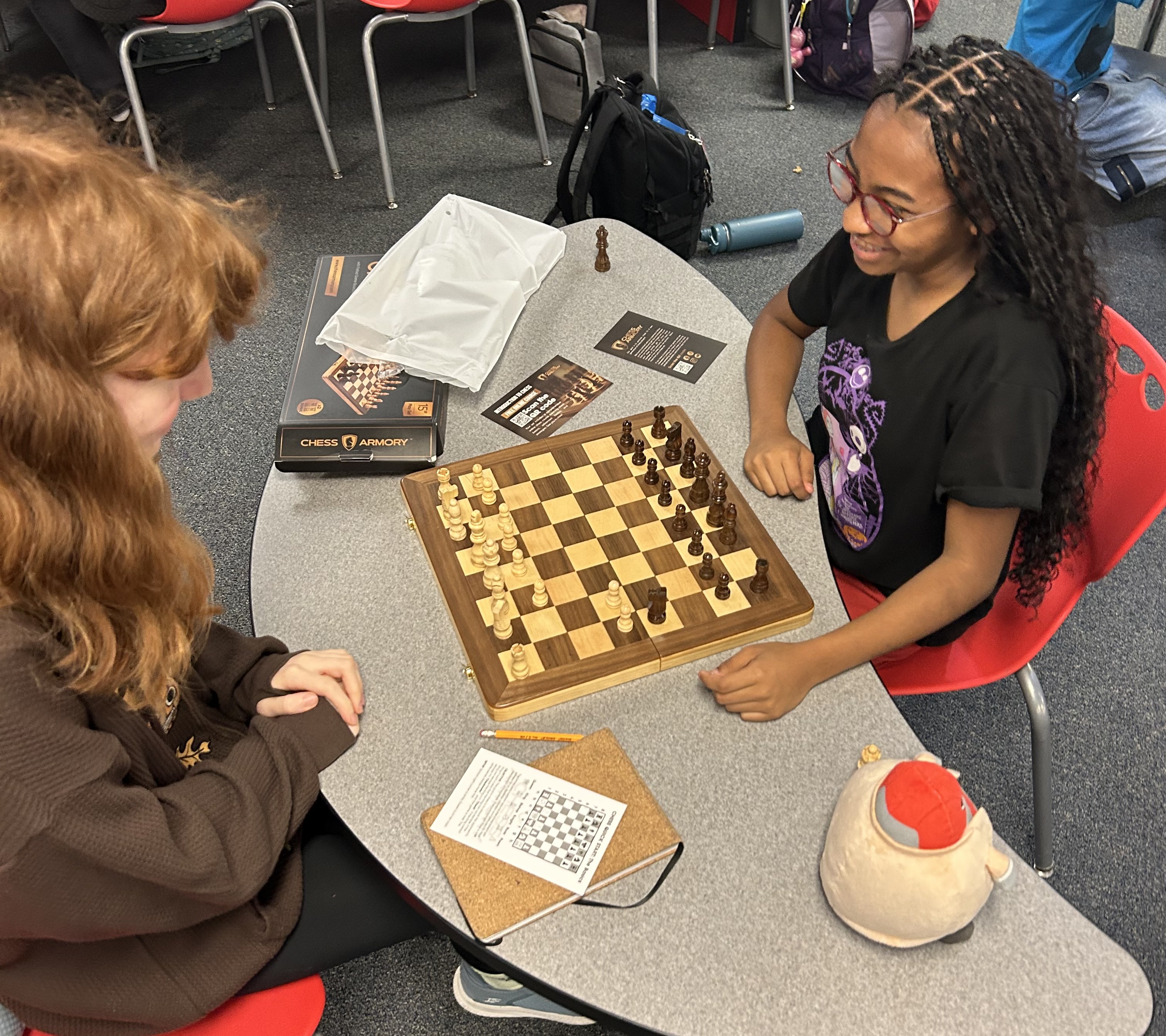 Two young women play chess. One wears glasses, the other a black T-shirt. A chessboard and pieces are on the table.