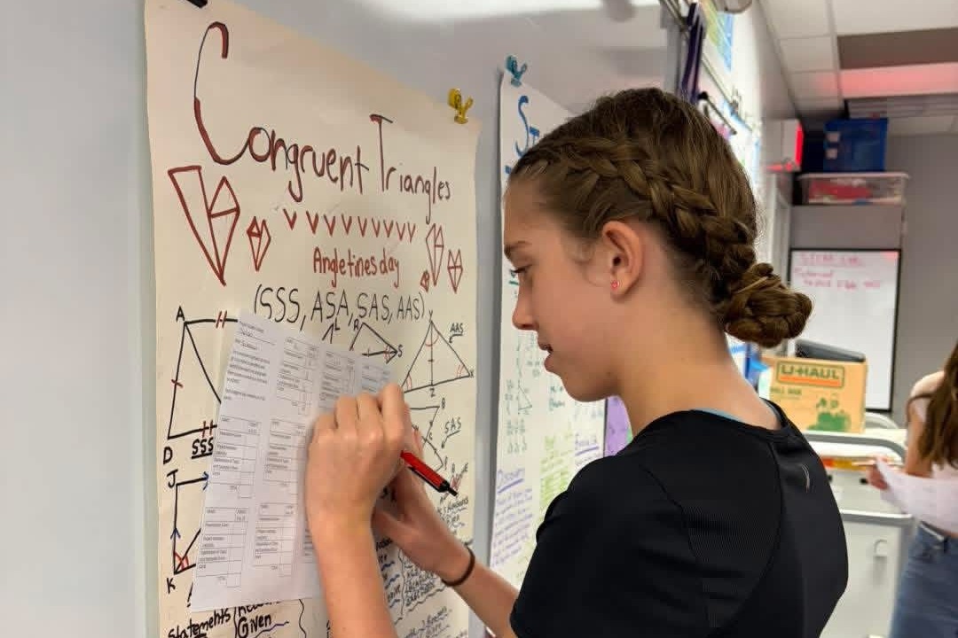 A student writes on a large paper in a classroom. The paper contains math terms and diagrams.