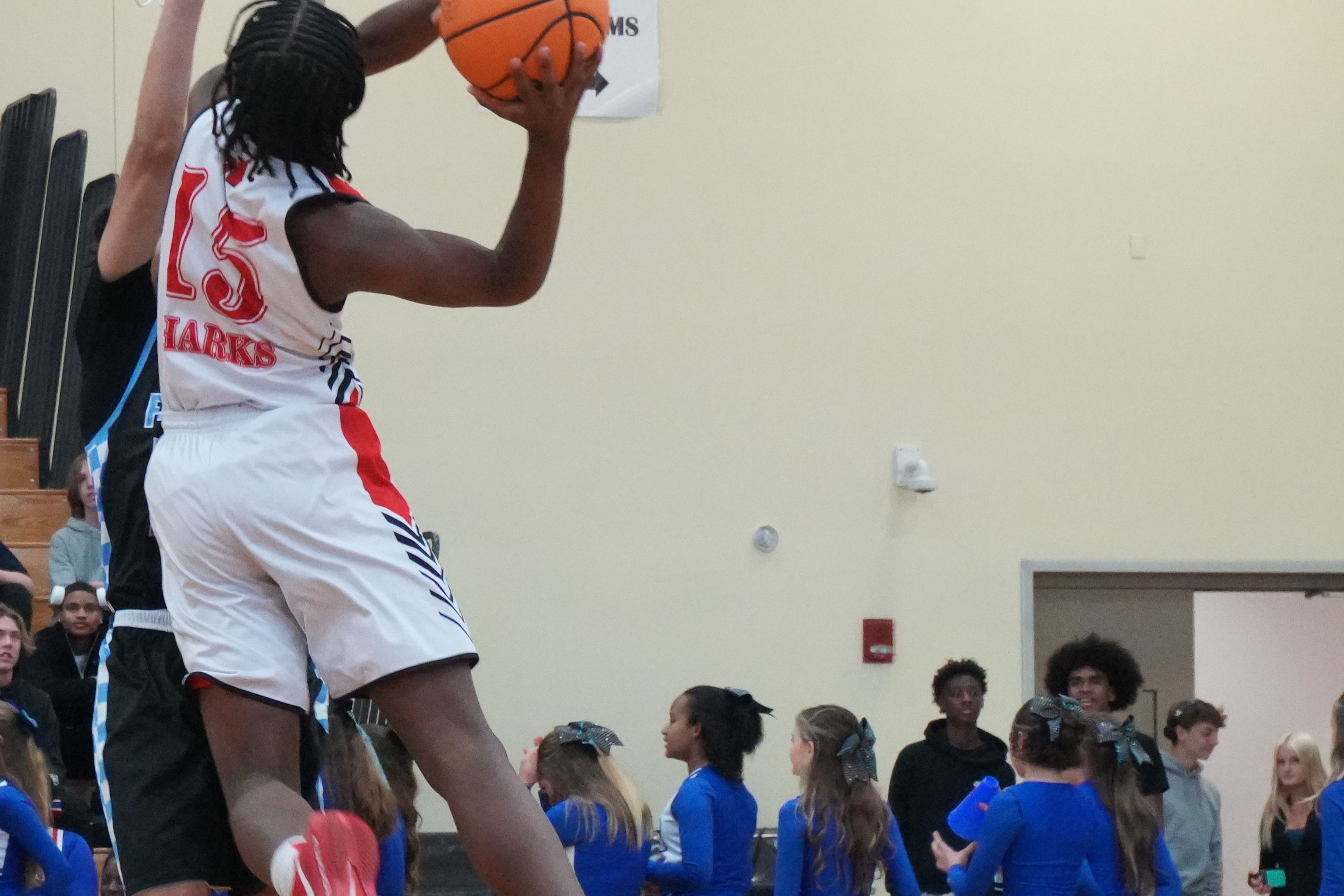 A player jumping to dunk a basketball while teammates in blue uniforms stand nearby. Background shows a gym with stairs and a group of spectators.