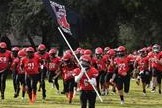 Team wearing red uniforms runs across a field, waving a flag. Green trees are in the background.