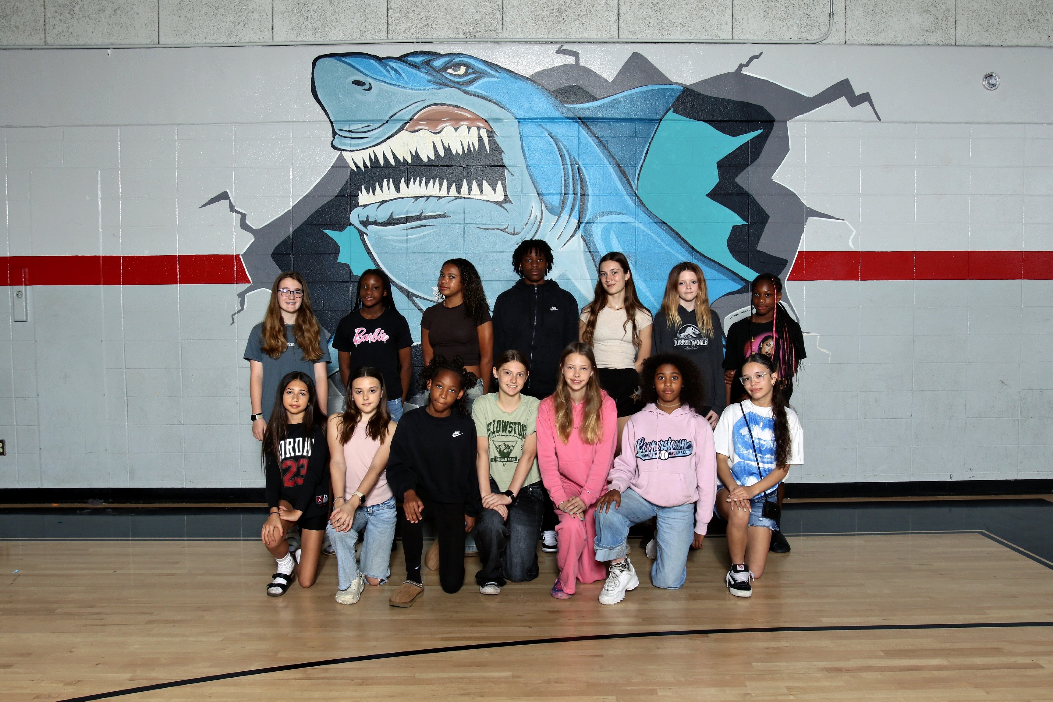 A group of people, likely a team, poses on a gym floor with a large shark mural in the background.