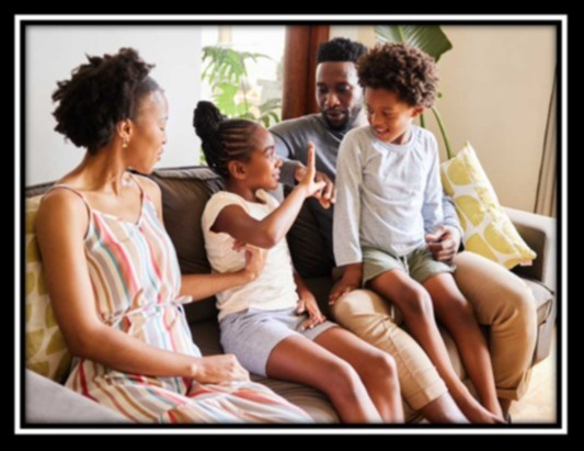 Four people sit on a couch; a woman in a dress, a man, and two kids. One child points and talks.