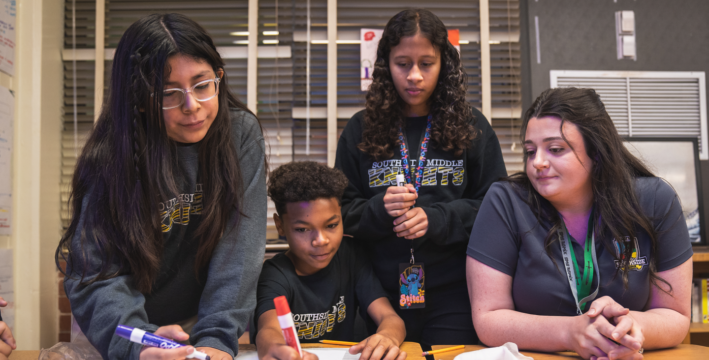 students at a table working on a project with their teacher's help